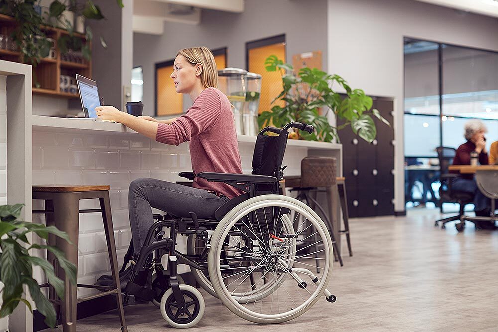 Das Bild zeigt eine Frau, die im Rollstuhl sitzt und in einer Cafeteria an einem Laptop arbeitet.